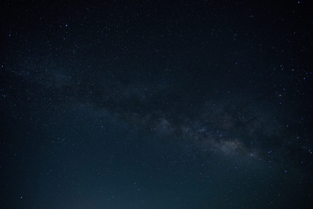 Under a clear night sky showcasing the Milky Way galaxy, the Universe unveils its splendor. Numerous stars are scattered across the dark expanse, while a dense, cloudy band runs diagonally, illustrating the galaxy's core. The lower part of this cosmic book has a subtle greenish hue.