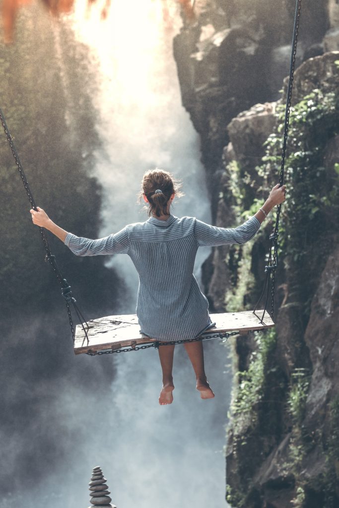 A person in a blue dress sits on a wooden swing, practicing mindfulness as they overlook a large waterfall. Suspended over the rocky gorge, with mist rising around them, the scene is both serene and adventurous, reminiscent of Tamme Buckner's meditative journeys.