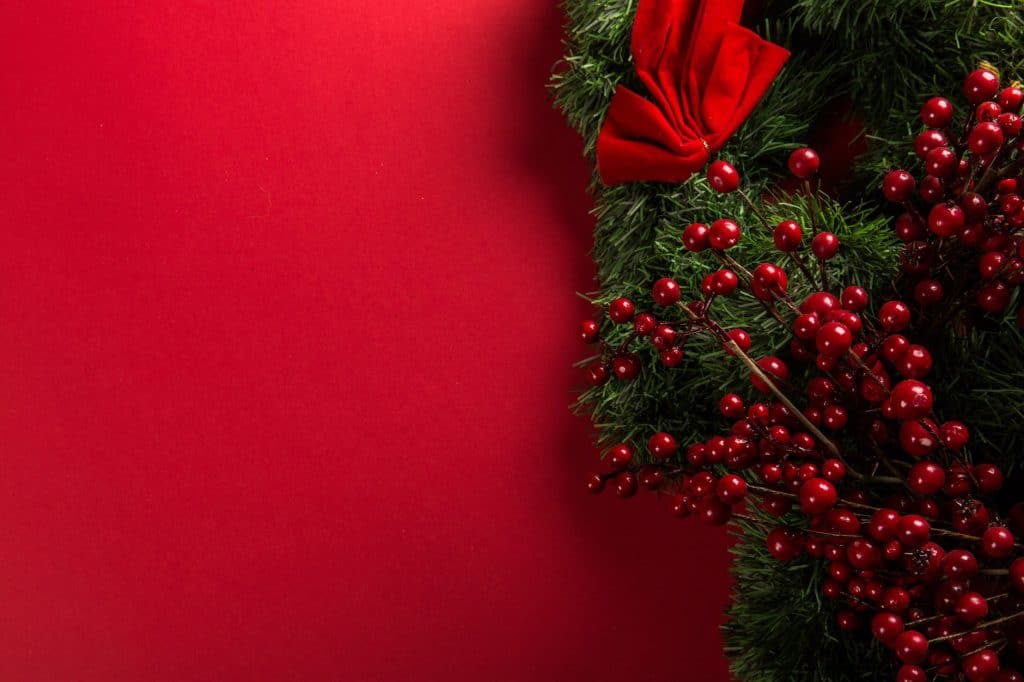 Christmas wreath, adorned with red berries and a bow on the right side against a solid red background, evokes festive joy. For those feeling overwhelmed, Tamme Buckner offers holiday stress tips to bring more peace during this cheerful season.