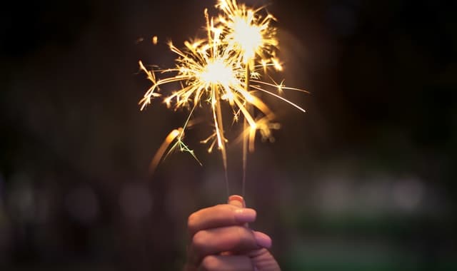 A hand holds two lit sparklers against a dark, blurred background, creating a warm glow with sparks radiating outward, reminiscent of New Year's resolutions igniting new beginnings.