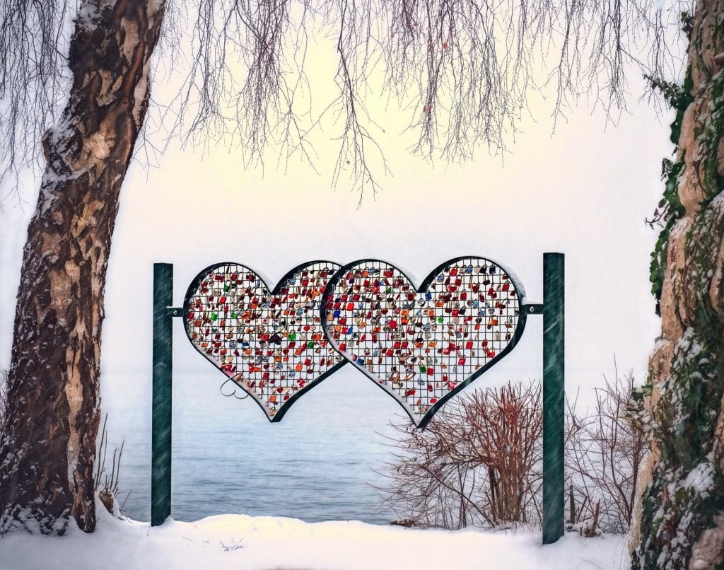 Two heart-shaped metal structures filled with colorful padlocks stand between two snow-covered trees, celebrating Valentine sentiments. The background reveals a serene, snowy landscape with a view of a calm body of water.