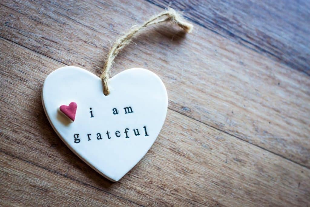 A heart-shaped ceramic ornament with a twine loop rests on a wooden surface. It reads "i am grateful" in black text, exuding genuine gratitude, with a small, red heart-shaped decoration attached. Inspired by Mariah Ehlert's heartfelt designs, this piece is both touching and sincere.