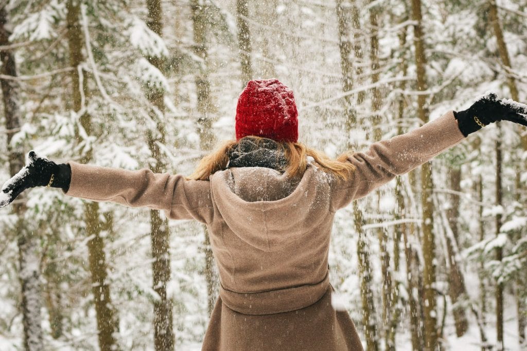 Stefanie Rothert, wearing a red knit hat, brown coat, and black gloves, stands in a snowy forest reminiscent of a scene from middle school art class. Arms outstretched, she looks up as snow falls around her. The trees, blanketed in white, add to the serene winter tableau.
