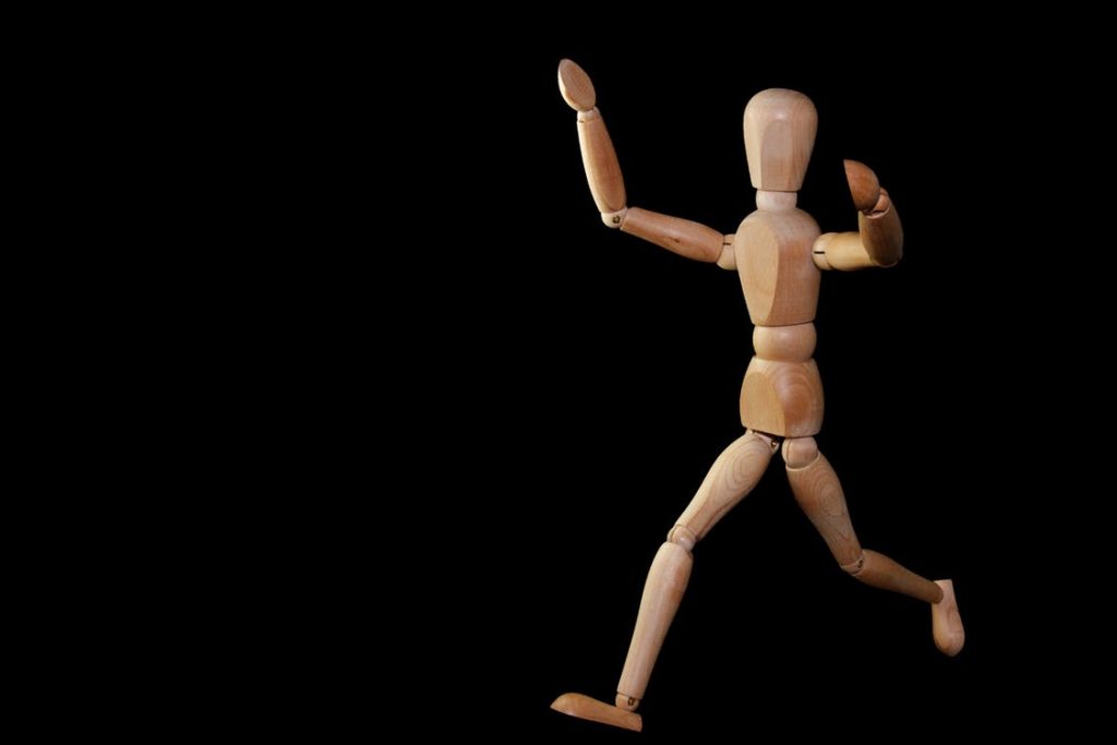 A wooden artist's mannequin, reminiscent of a Travis Rumsey masterpiece, is posed in a dynamic running position against a black background. Captured mid-stride with one arm raised and the other angled downward, it conveys the stress of motion and captures the essence of speed.