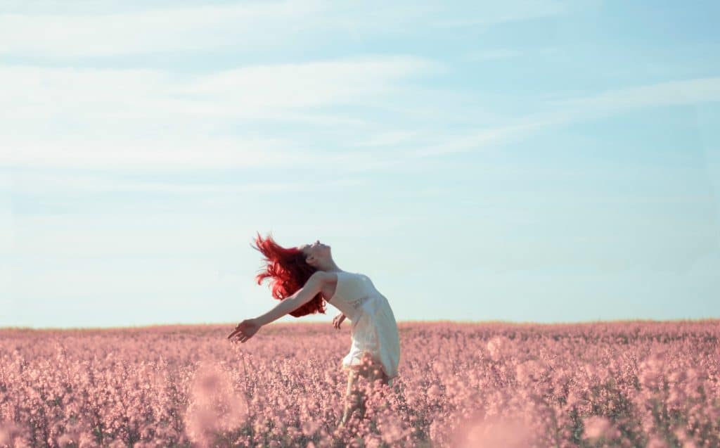 Danielle Rachlin, with long red hair, dances joyfully in a vast field of pink flowers under a clear blue sky. She wears a light dress, her arms outstretched as she leans back, embracing the natural surroundings and leaving stress far behind.