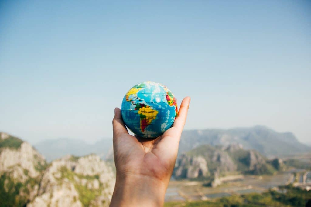 A hand holds a small globe against a mountainous landscape under a clear blue sky, capturing the essence of joy and exploration. The vibrant continents and oceans seem to speak a universal language of adventure and discovery.
