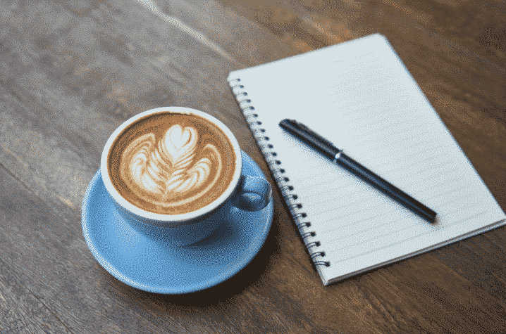 A cup of coffee with latte art in a blue saucer sits beside an open spiral notebook and a black pen on a wooden table, embodying Emily Geisler's approach to work-life balance. The latte art resembles a leaf pattern, inspiring focus and creativity.