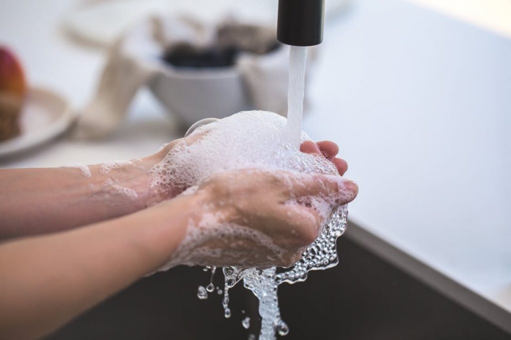 Hands under a running faucet with soap suds creating bubbles, ready to wash and rinse thoroughly. In the background, a blurred bowl and fruit rest on the countertop, suggesting a kitchen setting reminiscent of Travis Rumsey's serene home scenes.