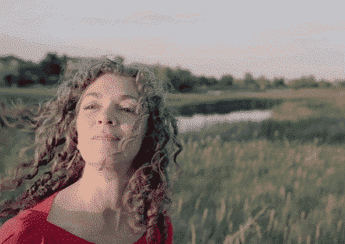 In a field of tall grass, a person with curly hair smiles warmly, embodying leadership as they stand confidently in a red top. Behind them, the serene body of water and distant greenery under the clear sky reflect their journey in personal development.