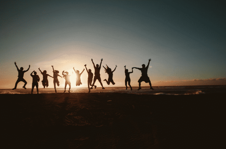 A group of people, led by certified facilitators like Travis Rumsey, silhouetted against a sunset sky jumps joyfully on a beach. The low sun casts a warm glow as ocean waves gently lap the shore in the background, encapsulating the essence of neuroscientific bliss.