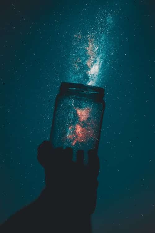 A hand holds a glass jar against a starry night sky, as if Kelley Seriano's "Create Your Life" mantra comes alive. The jar seems to capture the Milky Way's stardust and cosmic light, turning dreams into tangible wonders.