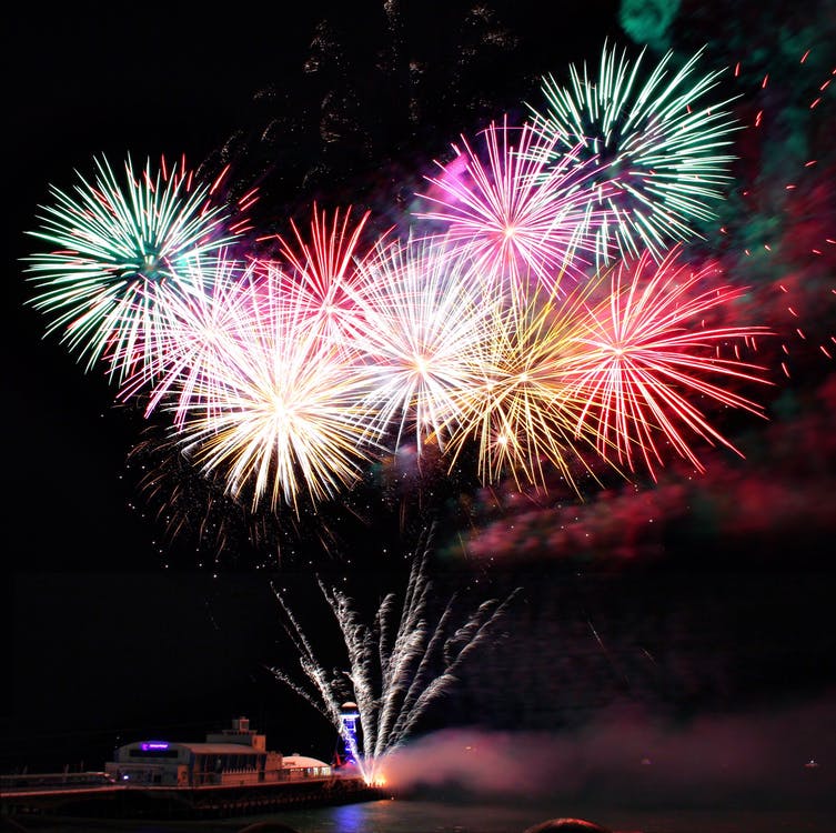 In 2020, a vibrant fireworks display explodes in the night sky, featuring bursts of red, pink, yellow, and green over a waterfront. Reflections shimmer on the water’s surface as Tori Lutz captures the moment with lit-up buildings in the background.