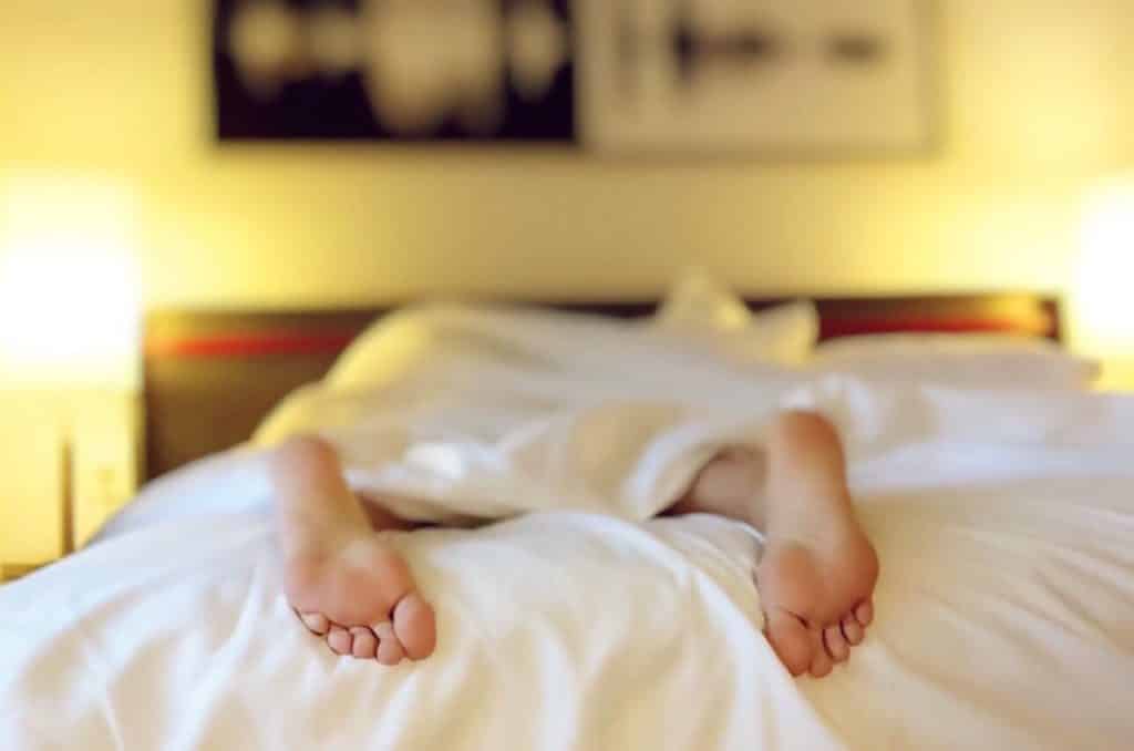 A person is lying in bed, covered by a white blanket, with only their bare feet visible, sticking out from the bottom. The room, softly lit by two bedside lamps and adorned with blurred artwork, offers a serene ambiance vital for mental health and restful sleep.