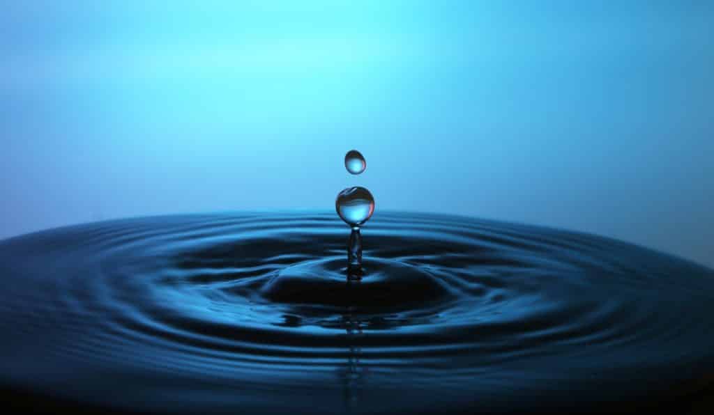 A water droplet falls into a pool, creating ripples on the surface—a moment of self-care captured in nature. The background is a gradient of blue shades, emphasizing the droplet in mid-air and the resulting splash.