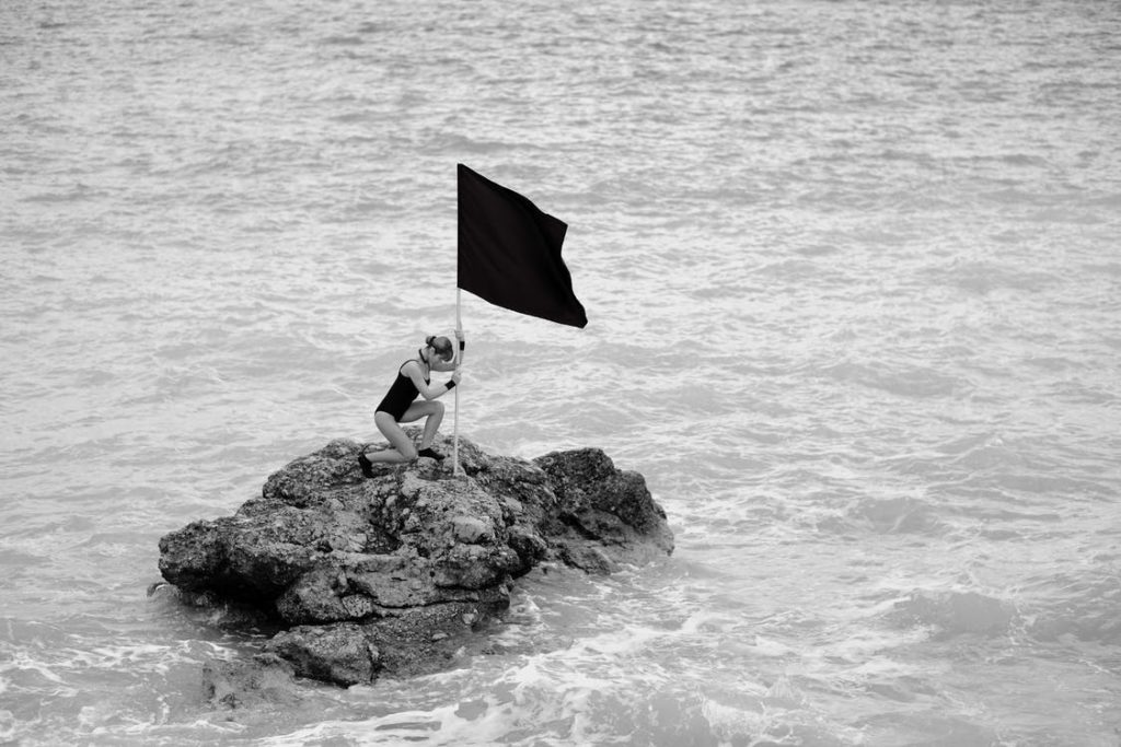 Kelley Seriano kneels on a small rocky outcrop in the sea, holding a large black flag. Wearing a swimsuit with a mask perched on their head, they embody vulnerability amidst the endless ocean. The scene unfolds in black and white, capturing an empowering connection with nature.