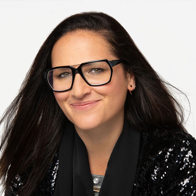 A smiling person exudes personal power, their long dark hair framing large black glasses. Dressed stylishly in a black sequined jacket and scarf, they stand against a plain white background, embodying a confident mindset.