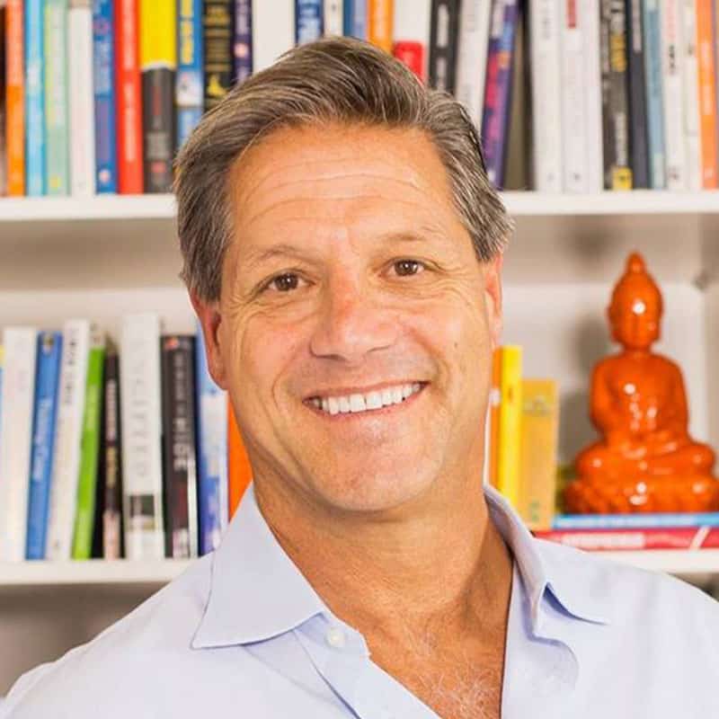 A man with short gray hair wearing a light blue shirt smiles confidently in front of a bookshelf filled with colorful books. A small orange statue of a Buddha reflects his mindful mindset, and the year 2020 stands out as the beginning of his journey toward personal power.