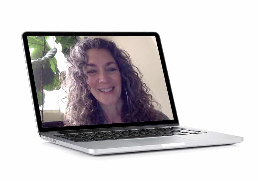 A woman with curly hair radiates personal power as she smiles during a video call on her laptop. The device sits on a white surface, with a green plant thriving in the background, reminding her to maintain a positive mindset.