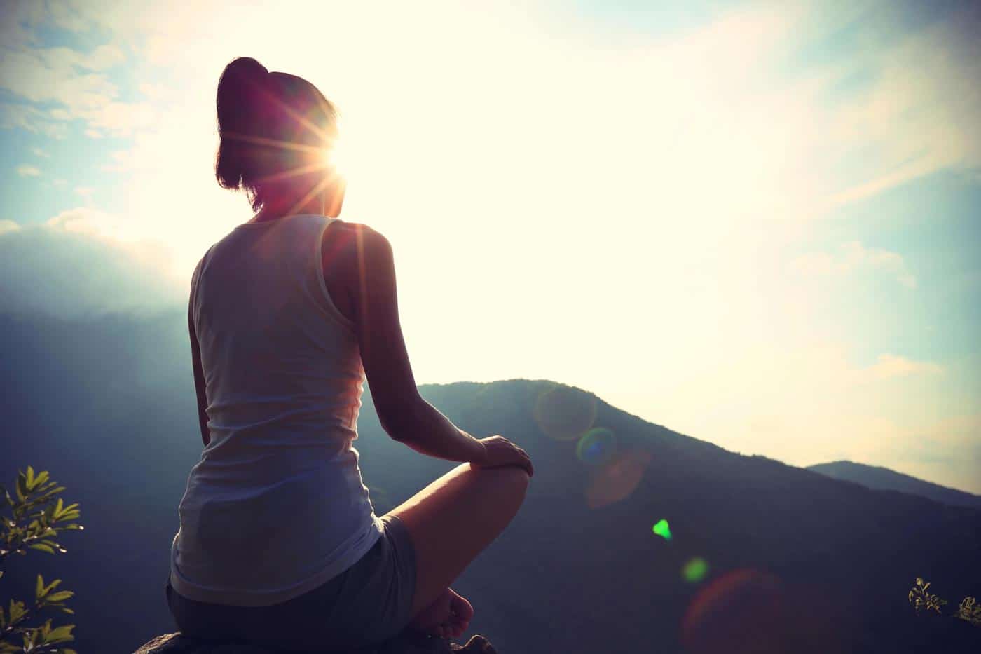 A person sits in a meditative pose, embracing their personal power and mindset, facing a scenic view of mountains with the sun shining brightly. They appear contemplative, silhouetted by the sunlight. The scene conveys tranquility and a deep connection with nature.