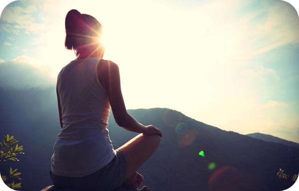 A person sitting on a rock, wearing a sleeveless top, is silhouetted against the bright sun. Embracing their personal power, they face a scenic view of mountains and clear skies in 2020, suggesting a tranquil, reflective moment in nature that inspires a powerful mindset.