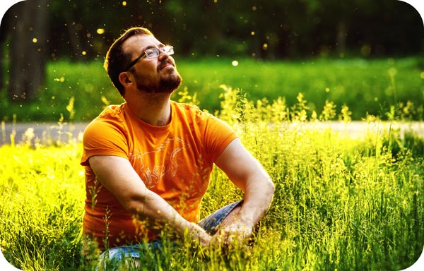 A man with a beard and glasses sits in a sunlit field, wearing an orange t-shirt and jeans. He gazes upward with a thoughtful expression, embodying a mindset of personal power amid the tall grass and small, floating particles that dance in the warm light.