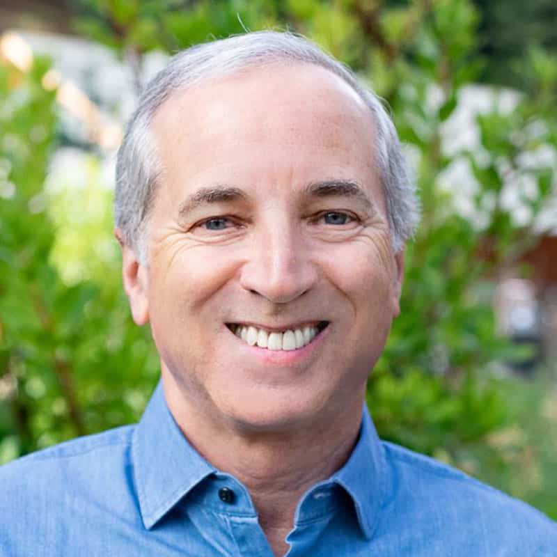 Smiling man with gray hair wearing a blue shirt, standing outdoors amidst green foliage, radiates a mindset of personal power.