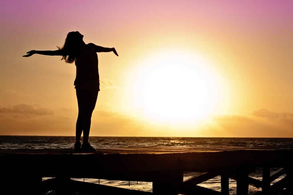 A silhouette of a person stands on the dock with arms outstretched, embracing the vibrant sunset that paints the ocean in shades of orange, pink, and purple. Channeling Kelley Seriano's essence, it's a moment of letting go as the sun sinks low on the horizon.
