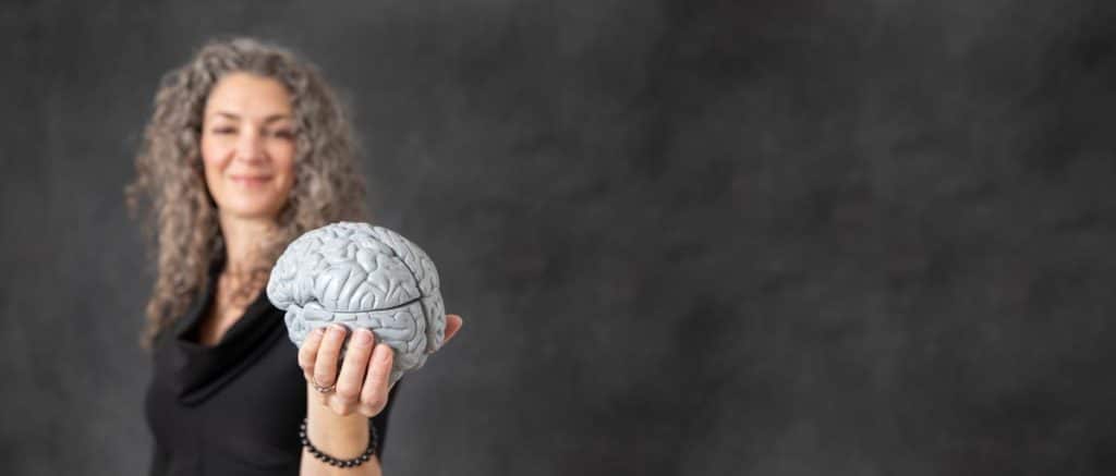 A woman with curly gray hair holds a model of a brain out towards the camera. She is wearing a black top and smiling slightly, with a blurred dark gray background.