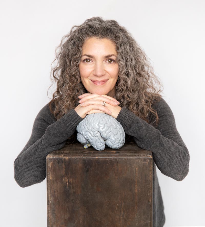 A person with curly gray hair, wearing a dark sweater, smiles gently, leaning on a dark wooden box. Their hands rest on a model of a brain placed on the box. They are set against a plain white background.