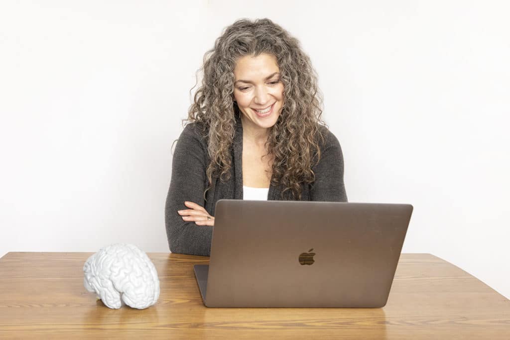 A woman with curly hair is sitting at a wooden table, smiling and looking at an open laptop. A brain model is placed on the table beside her. The background is a plain white wall.