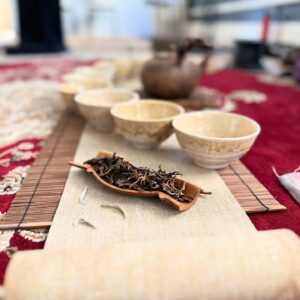A serene tea ceremony setup with four empty cups, a brown teapot, and a tray of loose tea leaves on a woven mat. The scene rests like an oasis on the red and beige carpet, enveloping you in tradition and tranquility.
