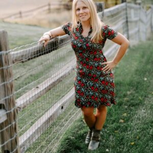 A woman with long blonde hair stands by a wire and wooden fence on a grassy lawn, wearing a black dress with a red and green floral pattern and ankle boots. Trees and a field are visible in the background.