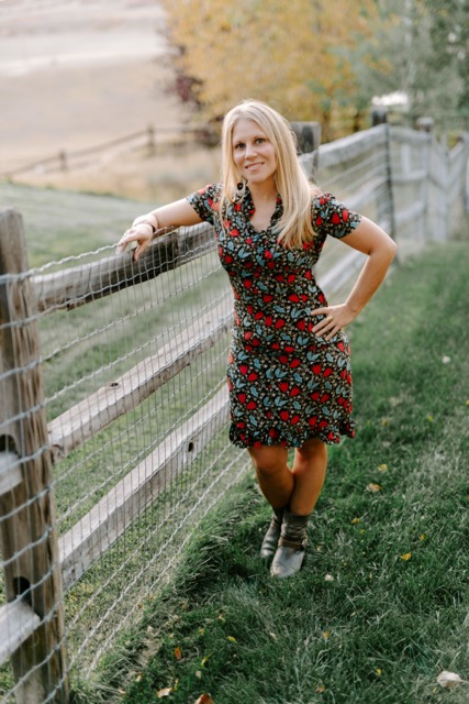 A woman with long blonde hair stands by a wire and wooden fence on a grassy lawn, wearing a black dress with a red and green floral pattern and ankle boots. Trees and a field are visible in the background.