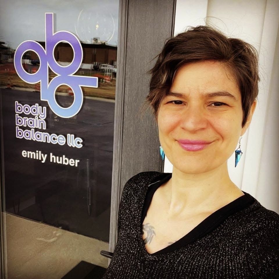 A person with short brown hair and blue earrings smiles in front of a glass door displaying the logo and name "body brain balance llc" and "emily huber." The background shows a reflection of a street and buildings.