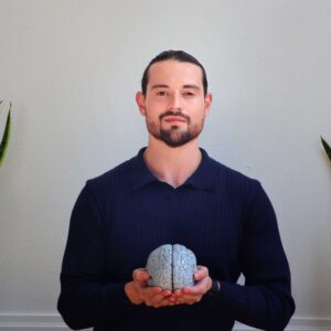 A man with dark hair and a beard, wearing a navy sweater, stands indoors holding a model of a human brain. He is flanked by two snake plants against a plain light-colored wall.