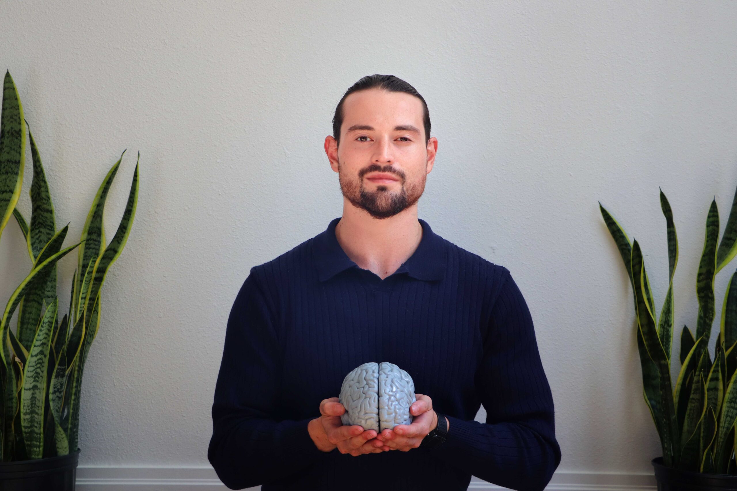 A man with dark hair and a beard, wearing a navy sweater, stands indoors holding a model of a human brain. He is flanked by two snake plants against a plain light-colored wall.