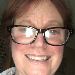 A woman with short, reddish hair and large, dark-rimmed glasses smiles at the camera in a softly lit indoor setting.