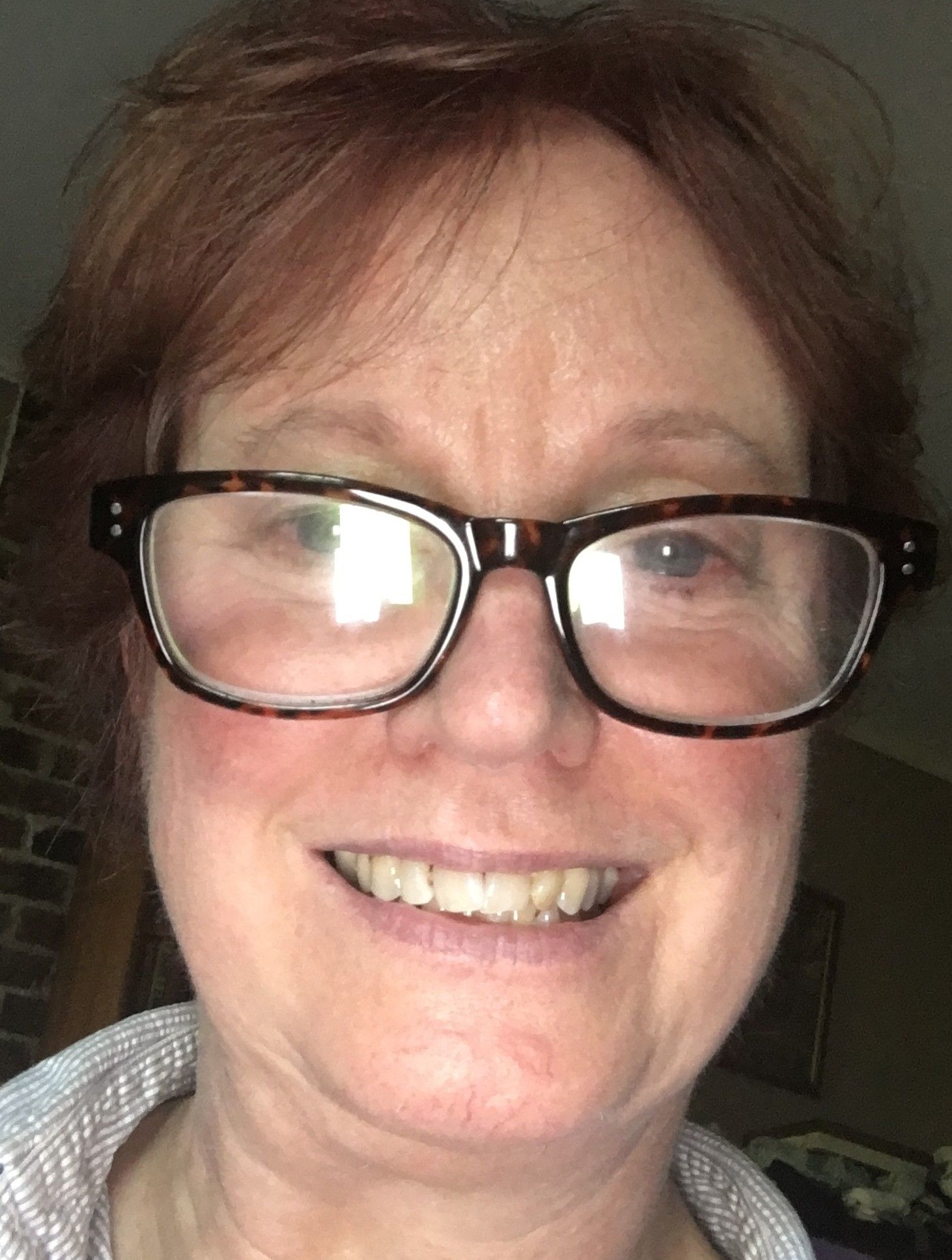 A woman with short, reddish hair and large, dark-rimmed glasses smiles at the camera in a softly lit indoor setting.