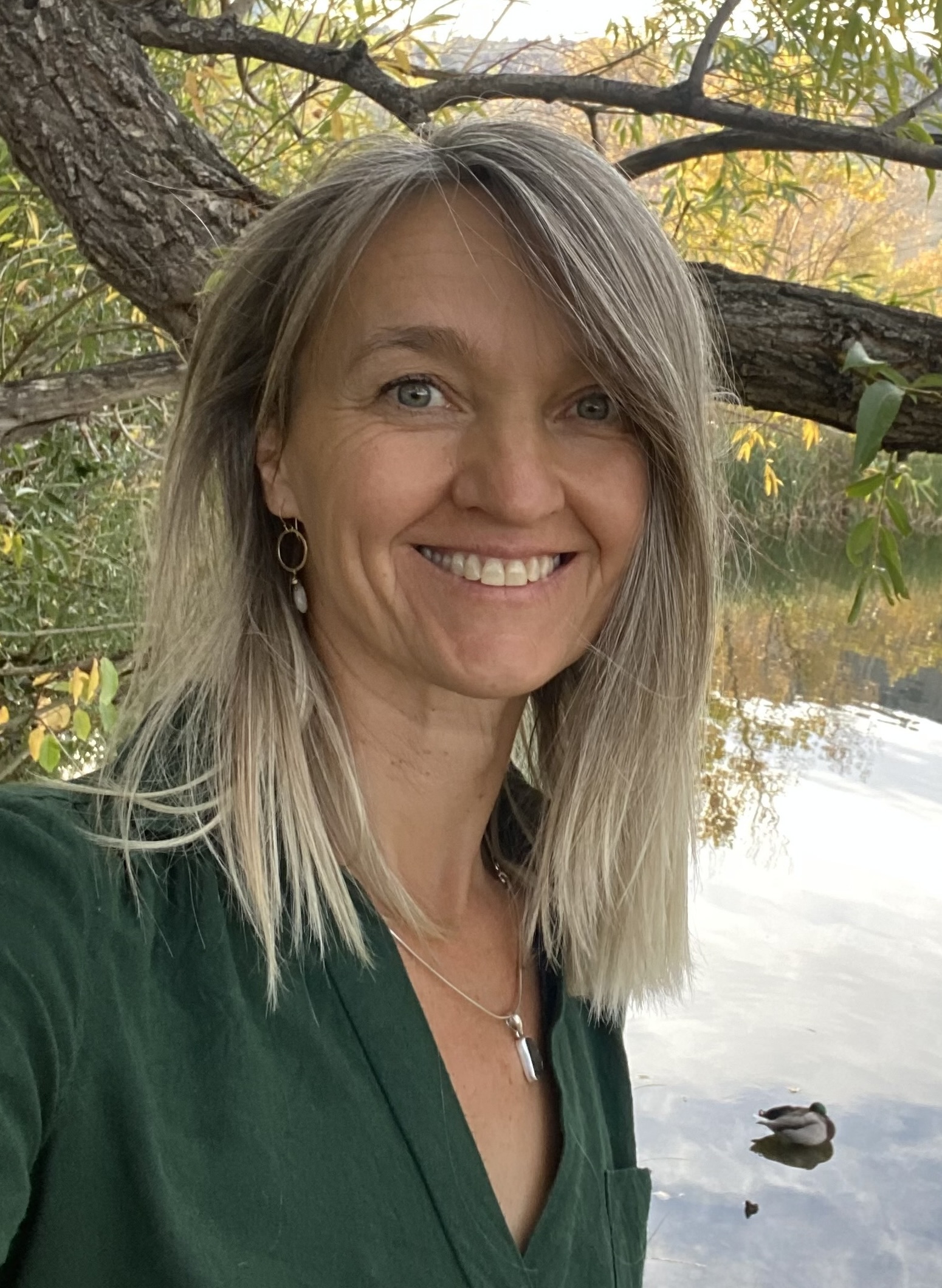 A woman with blonde hair and a green top smiles outdoors near a tree and a calm body of water with a duck in the background.