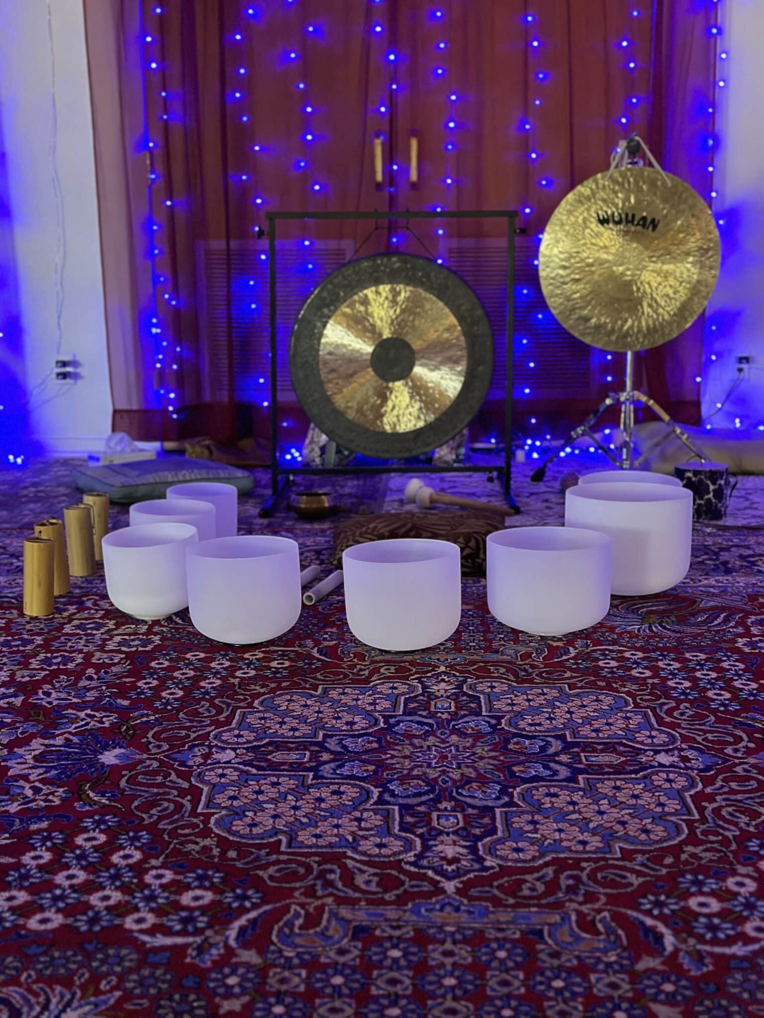 A collection of white crystal singing bowls is arranged on a patterned carpet, setting the scene for a serene sound bath. A large gong with a stand and another mounted gong complement the ambiance, while blue string lights add a soothing touch to this perfect setting for sound therapy.