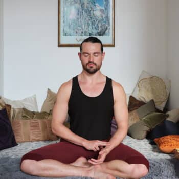Anthony Sosa, an LPCC expert, sits cross-legged on a cushion in a meditative pose, wearing a black tank top and burgundy pants. The room is adorned with vibrant cushions and a framed picture behind him. Eyes closed and hands resting on knees, he immerses himself in his breathwork practice.