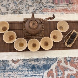 A textured clay teapot and six small tea bowls arranged in a semi-circle on a bamboo mat, creating a serene tea house setting with a small tray of loose-leaf tea and a single green leaf to the side.
