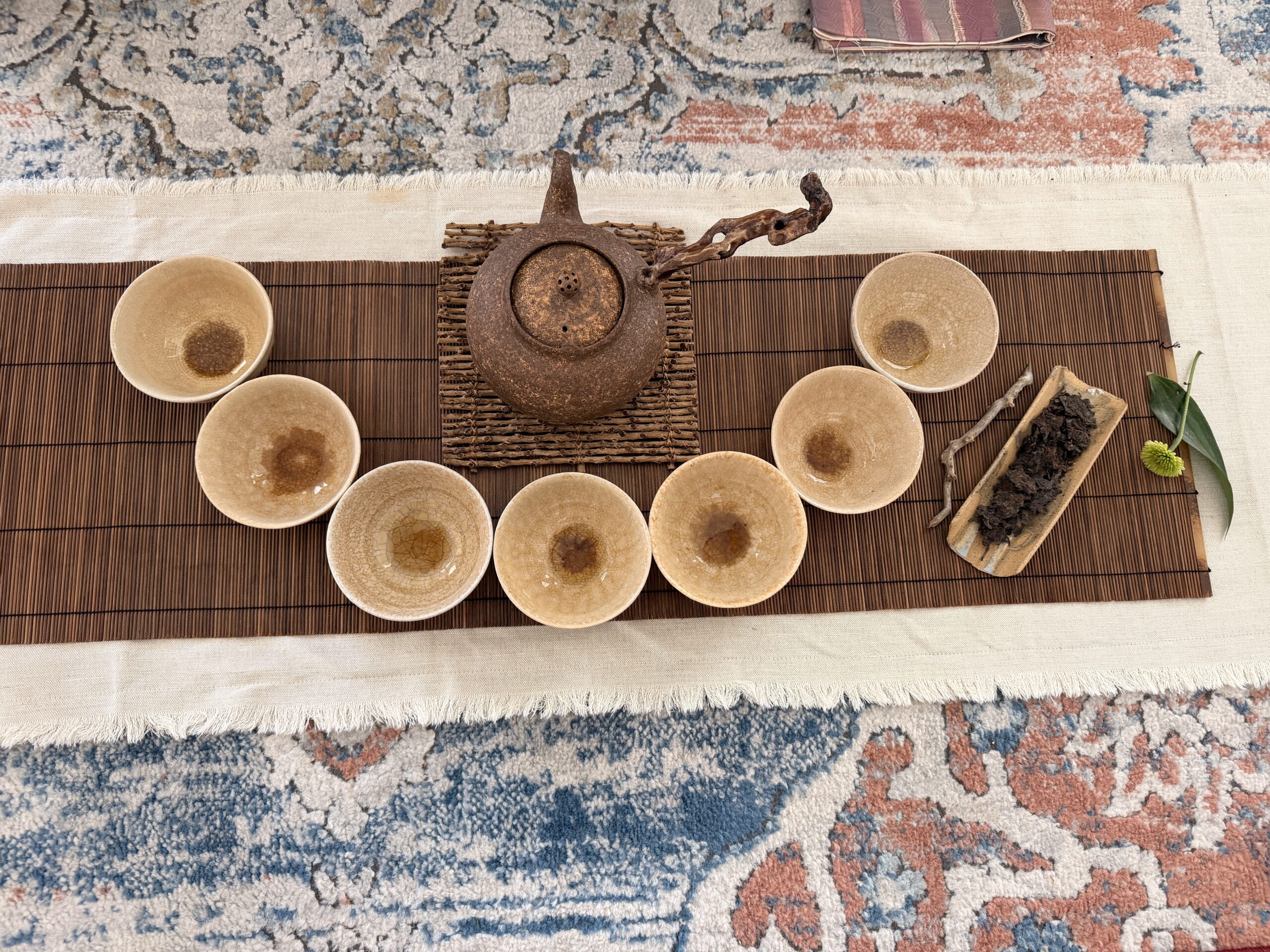 A textured clay teapot and six small tea bowls arranged in a semi-circle on a bamboo mat, creating a serene tea house setting with a small tray of loose-leaf tea and a single green leaf to the side.