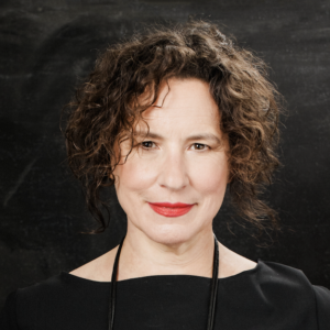 Woman with curly brown hair and red lipstick, wearing a black top and black necklace, posing in front of a dark background.