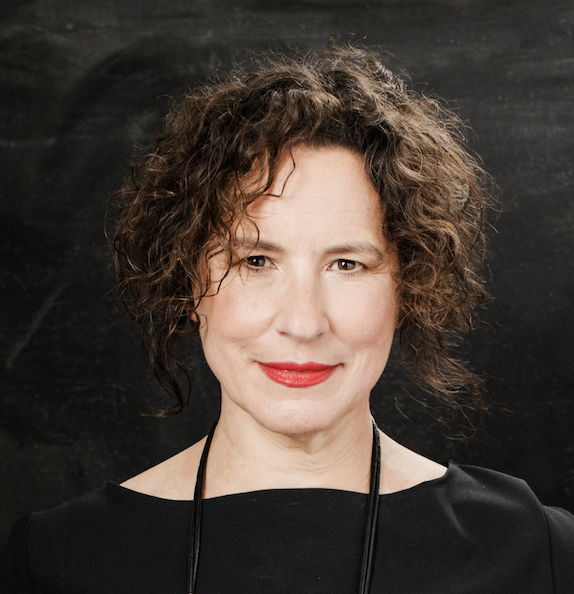 Woman with curly brown hair and red lipstick, wearing a black top and black necklace, posing in front of a dark background.