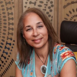 A woman with light brown hair and a patterned blue top smiles while seated indoors. A circular patterned wooden screen serves as the backdrop. She wears a pendant necklace.