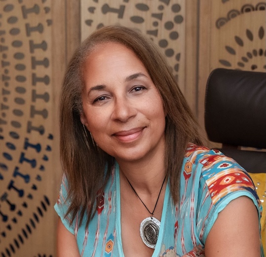 A woman with light brown hair and a patterned blue top smiles while seated indoors. A circular patterned wooden screen serves as the backdrop. She wears a pendant necklace.