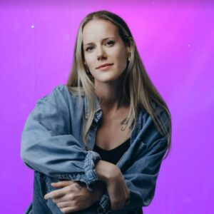 A woman with long blonde hair wearing a denim shirt and black top sits with her arms resting on her knee, against a vibrant purple background.