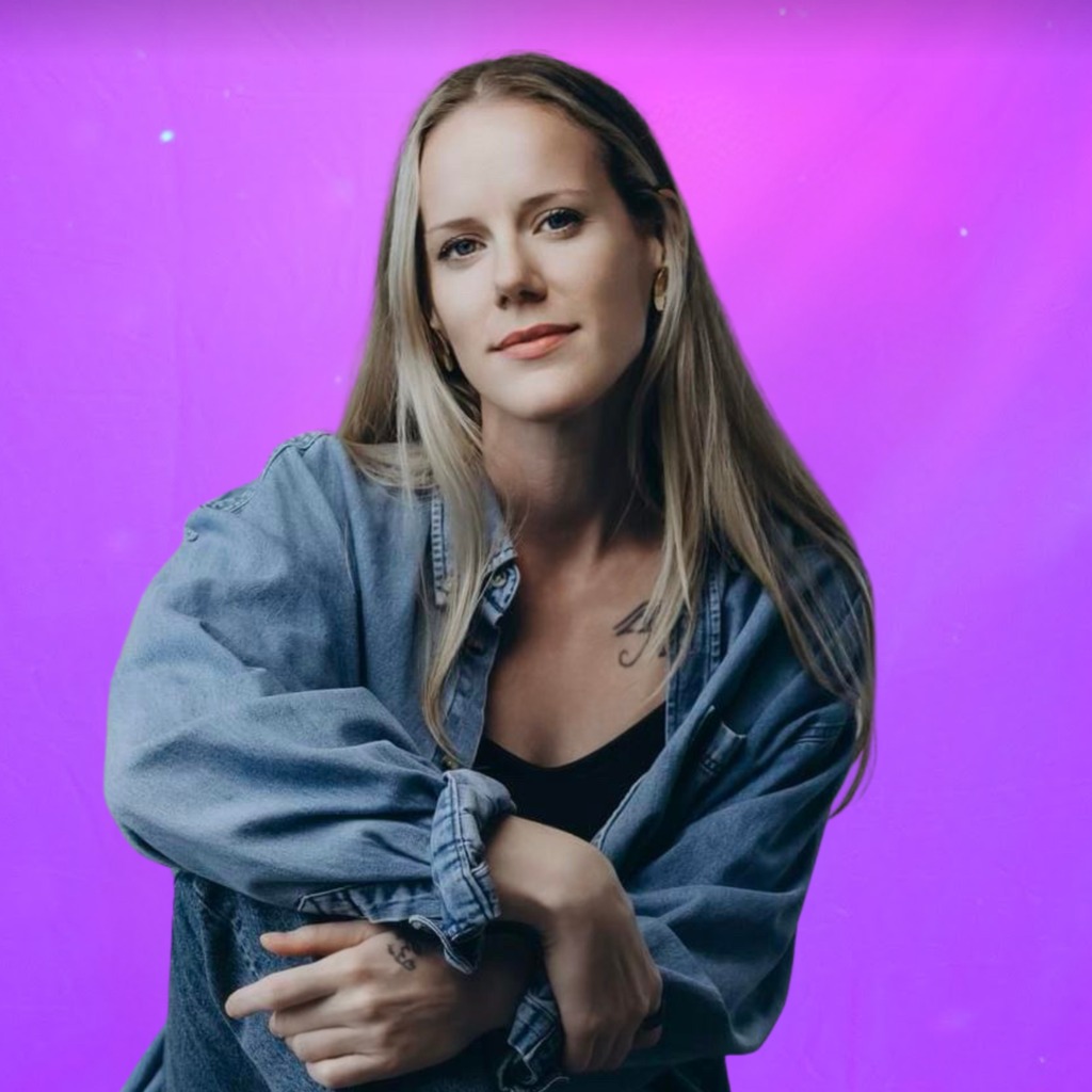 A woman with long blonde hair wearing a denim shirt and black top sits with her arms resting on her knee, against a vibrant purple background.