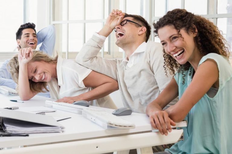 Four people sitting at a table with laptops, laughing joyfully. They appear to be in a bright office environment with large windows in the background. Their body language suggests shared amusement or enjoyment.